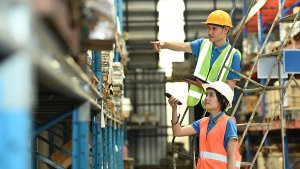 Male and female workers wearing hardhats and reflective jackets checking inventory boxes with barcode scanner in warehouse